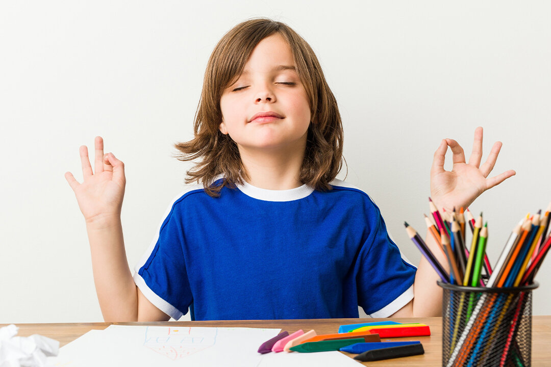 Kind sitzt mit geschlossenen Augen am Tisch und meditiert, daneben liegen bunte Stifte und Papier