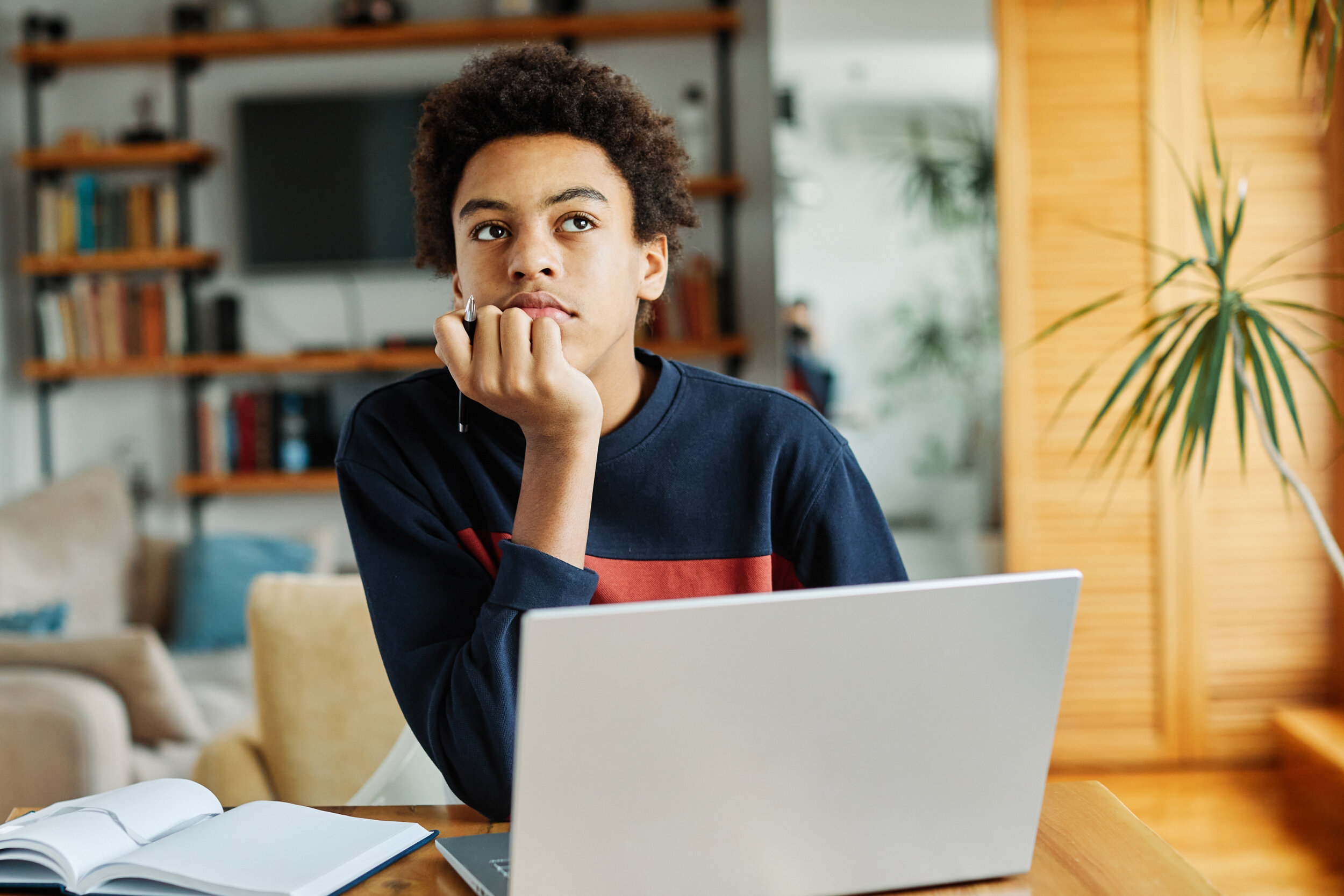  Nachdenklicher Schüler mit Laptop und Buch beim Wissencheck zuhause