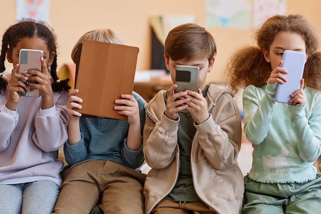 Vier Kinder sitzen nebeneinander und halten Smartphones oder ein Tablet vor das Gesicht.