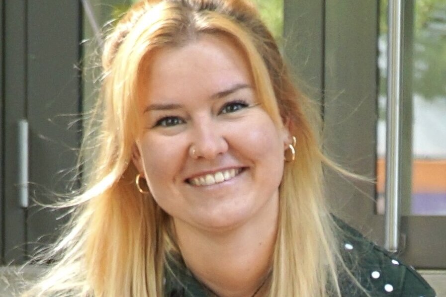 Melanie Lachmann, Redakteurin, Porträt mit warmem Lächeln und direktem Blick – lange rotgoldblonde Haare mit Wellen bis zur Schultermitte, blaugraue Augen, dunkelgrüne Bluse mit weißem Polka-Dot-Muster, goldene Ohrringe, sitzt entspannt mit gefalteten Händen, vertrauenswürdig und nahbar, moderne Außenkulisse mit grüner Vegetation im Hintergrund