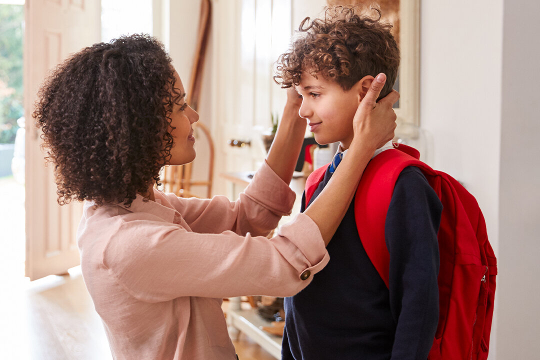 Mutter richtet ihrem Sohn mit Schulrucksack im Eingangsbereich liebevoll die Haare, bevor er das Haus verlässt.