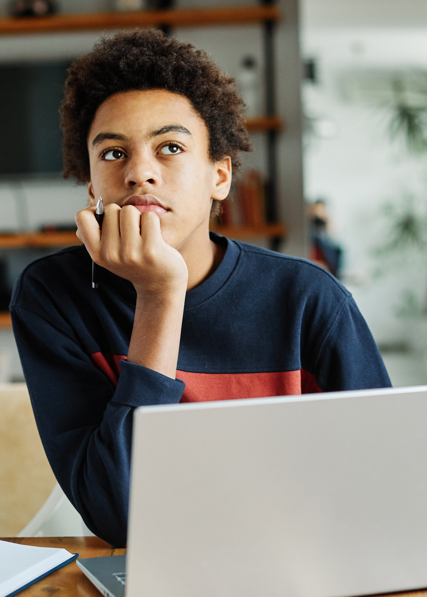 Jugendliche Person mit Afro-Frisur nachdenklich am Laptop, Hand am Kinn, Notizbuch und Stift, konzentriertes Lernen