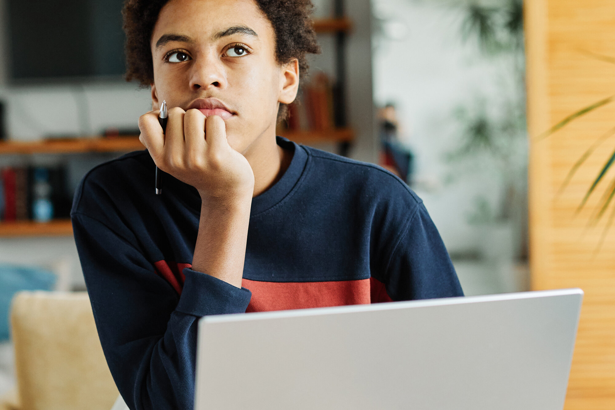 Jugendliche Person mit Afro-Frisur nachdenklich am Laptop, Hand am Kinn, Notizbuch und Stift, konzentriertes Lernen