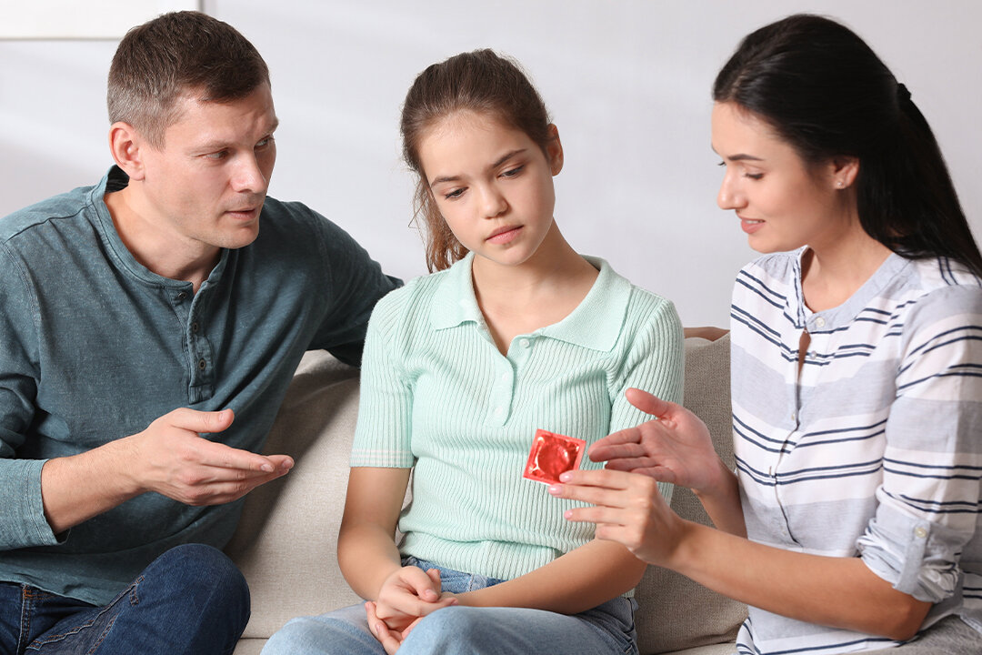 Zwei Erwachsene sprechen mit einem Mädchen auf dem Sofa und erklären ihr anhand eines Kondoms das Thema Verhütung