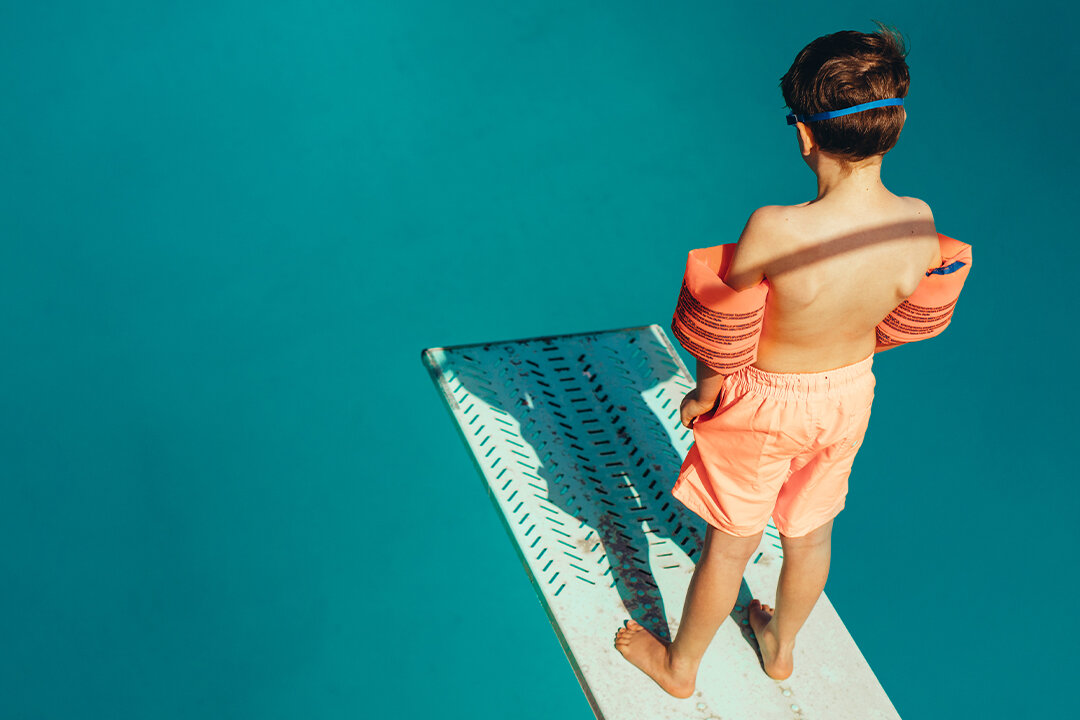 Junge mit Schwimmflügeln steht auf Sprungbrett im Schwimmbad und schaut ins Becken.