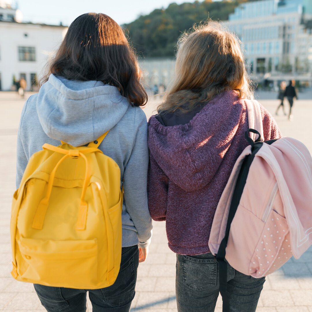 3 Teenager laufen über einen Platz. Man sieht sie von hintern. Sie tragen Rucksäcke.