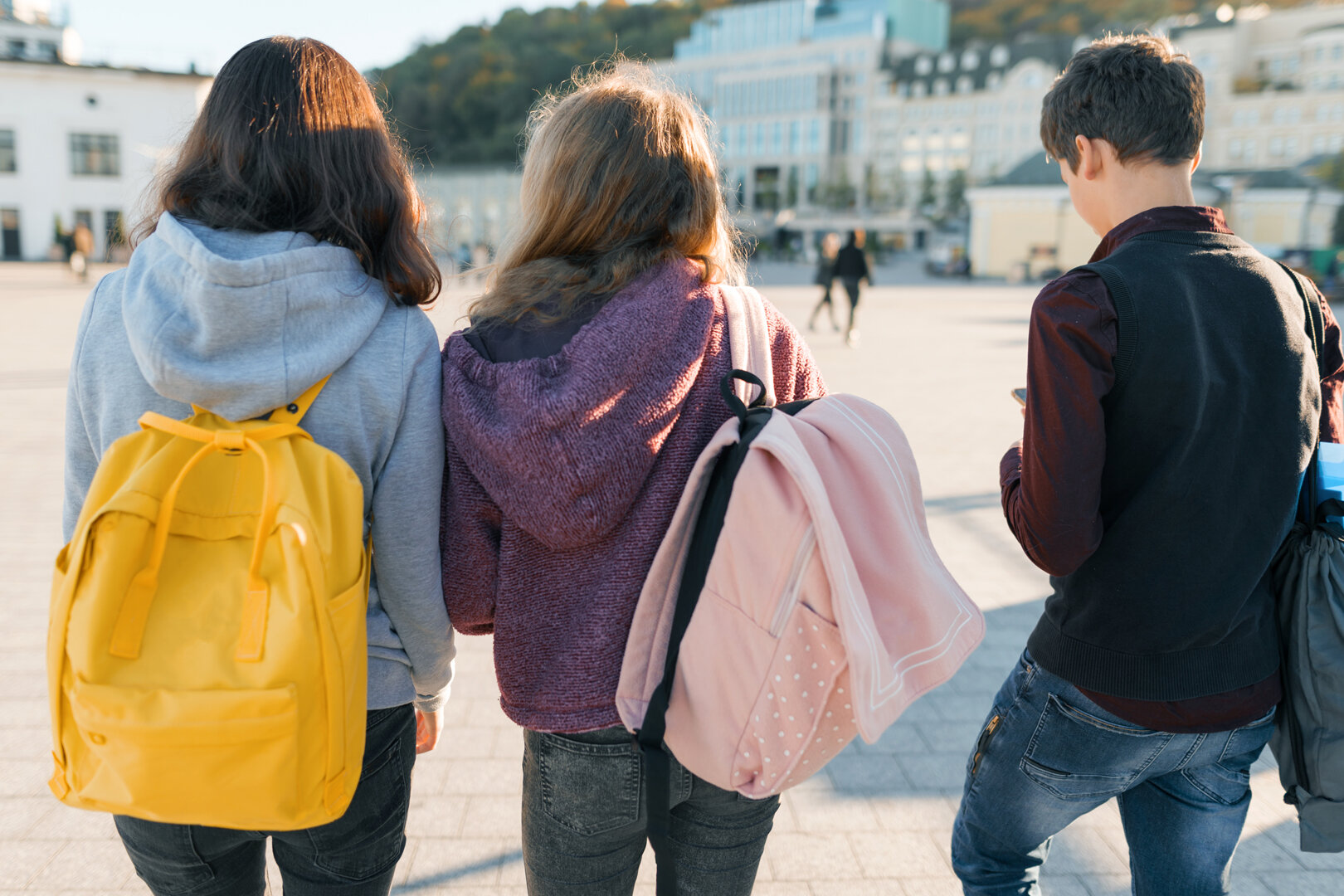 3 Teenager laufen über einen Platz. Man sieht sie von hintern. Sie tragen Rucksäcke.