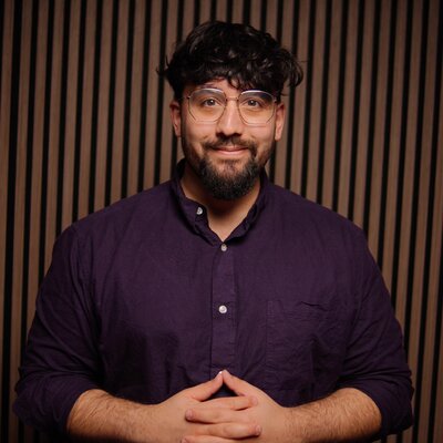 Lukas Maher, Redakteur, Porträt mit aufrichtigem Lächeln – dunkle, voluminöse Haare mit Textur, gepflegter dunkler Bart, klare Brille mit modernem Design, dunkles Navy-Hemd mit weißen Perlmuttknöpfen, Hände klassisch gefaltet, intellektuell und zugänglich, Vertrauen und Kompetenz