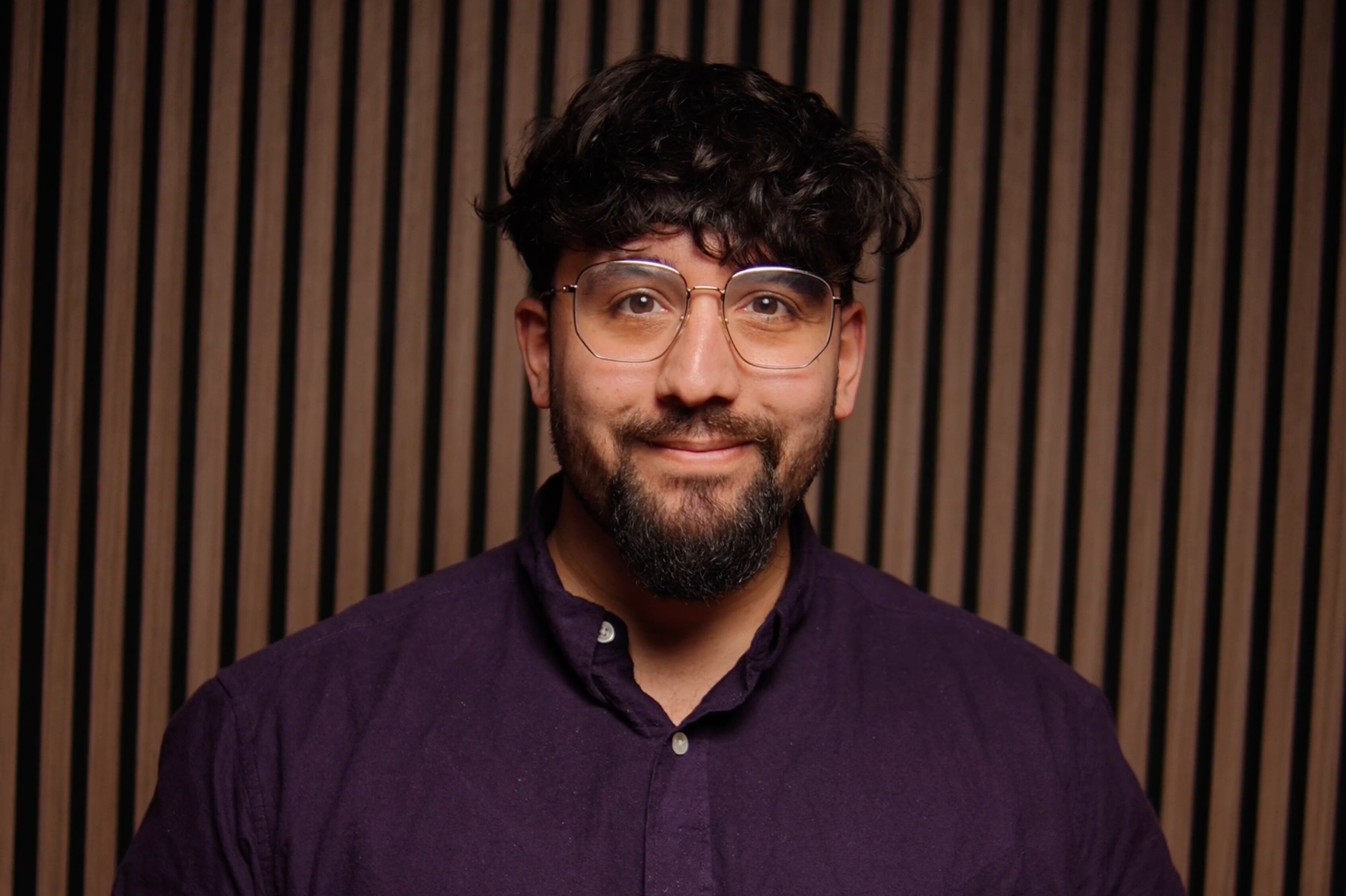 Lukas Maher, Redakteur, Porträt mit aufrichtigem Lächeln – dunkle, voluminöse Haare mit Textur, gepflegter dunkler Bart, klare Brille mit modernem Design, dunkles Navy-Hemd mit weißen Perlmuttknöpfen, Hände klassisch gefaltet, intellektuell und zugänglich, Vertrauen und Kompetenz