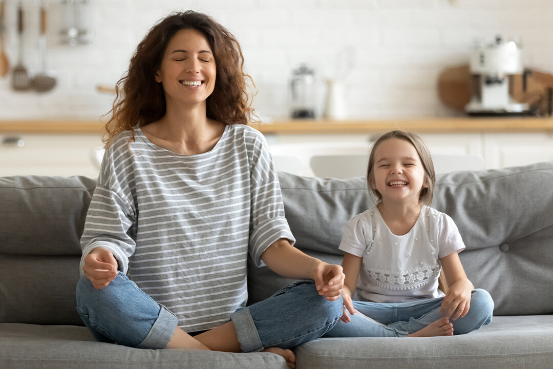 Mutter und Tochter praktizieren gemeinsam Achtsamkeit und Meditation zu Hause – beide sitzen entspannt im Schneidersitz auf dem Sofa mit geschlossenen Augen