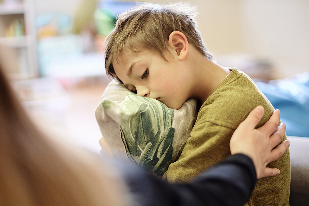 Ein Junge sitzt traurig auf einem Sofa und hält ein Kissen im Arm. Eine erwachsene Person legt ihm tröstend die Hand auf die Schulter.