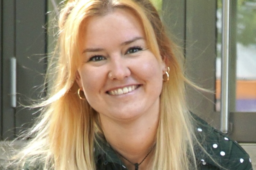 Melanie Lachmann, Redakteurin, Porträt mit warmem Lächeln und direktem Blick – lange rotgoldblonde Haare mit Wellen bis zur Schultermitte, blaugraue Augen, dunkelgrüne Bluse mit weißem Polka-Dot-Muster, goldene Ohrringe, sitzt entspannt mit gefalteten Händen, vertrauenswürdig und nahbar, moderne Außenkulisse mit grüner Vegetation im Hintergrund