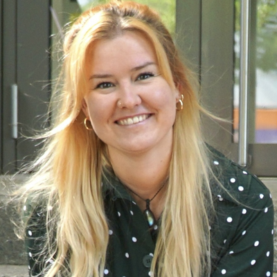 Melanie Lachmann, Redakteurin, Porträt mit warmem Lächeln und direktem Blick – lange rotgoldblonde Haare mit Wellen bis zur Schultermitte, blaugraue Augen, dunkelgrüne Bluse mit weißem Polka-Dot-Muster, goldene Ohrringe, sitzt entspannt mit gefalteten Händen, vertrauenswürdig und nahbar, moderne Außenkulisse mit grüner Vegetation im Hintergrund