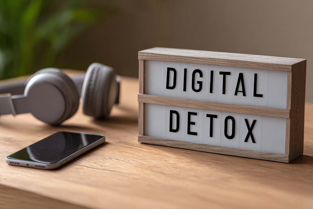 Leuchtbox mit Aufschrift „Digital Detox" auf Holzoberfläche, daneben abgelegtes Smartphone und Kopfhörer – Symbol für digitale Auszeit im Familienalltag