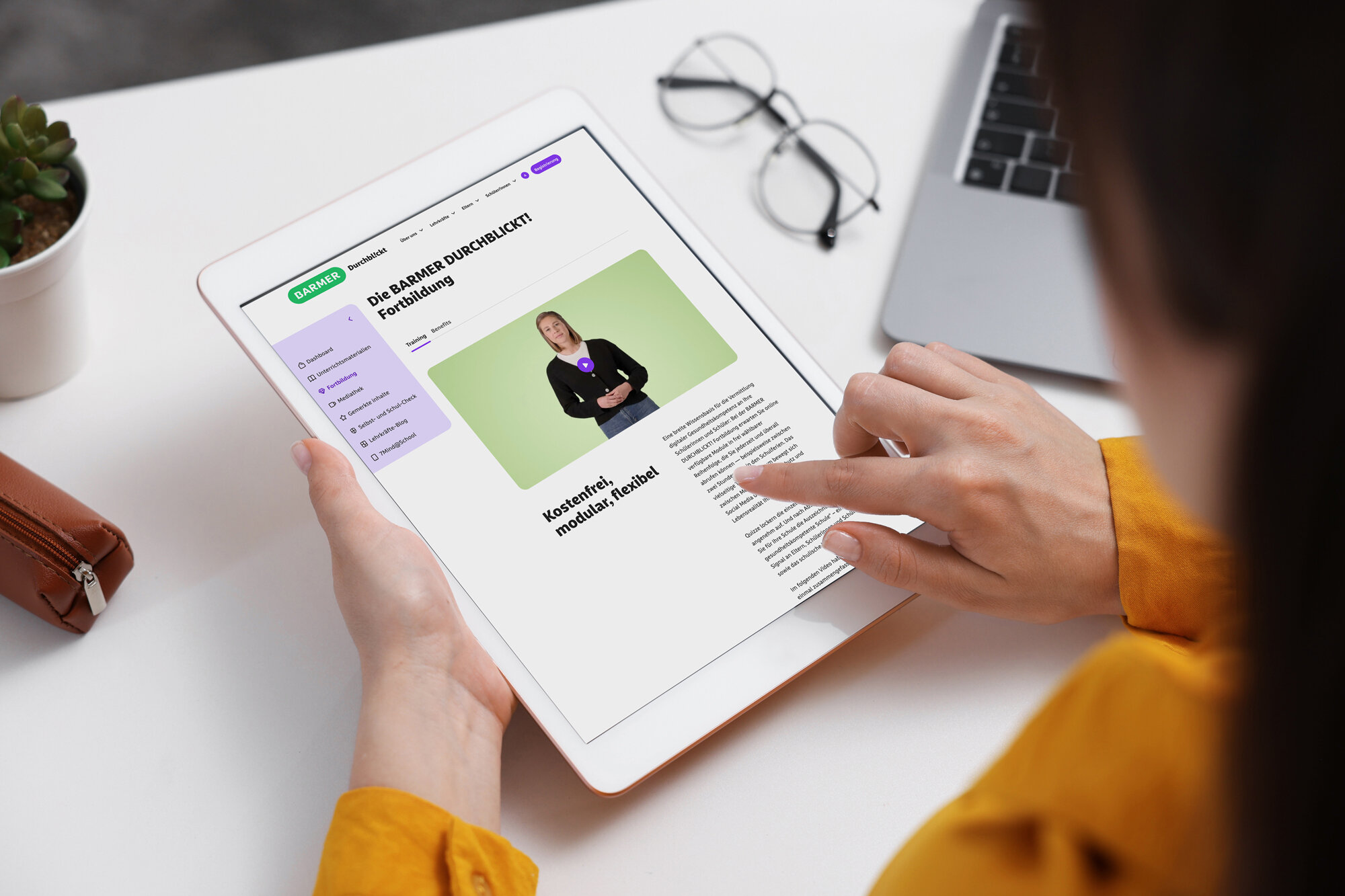 Mittelalte Lehrkraft mit dunkelbraunen Haaren in gelbem Pullover studiert konzentriert die BARMER Durchblickt Fortbildung auf Tablet am minimalistischen Schreibtisch – digitale Weiterbildung für Pädagogen