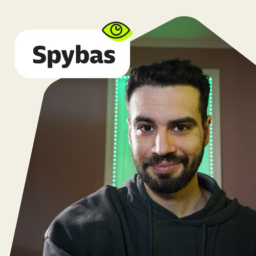 Porträt von Spyridon (Spybas), Gesicht verpixelt, dunkler Hoodie, grüne LED-Lichter, Label „Spybas“.