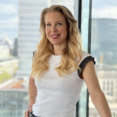 Viola Herrmann, Redakteurin, Porträt mit strahlendem Lächeln und direktem Blick – schulterlange goldblonde Haare mit Wellen, blaue Augen, leuchtend pinker Lippenstift, weißes T-Shirt, schwarzer Gürtel mit goldenem Schnalle, schwarzes Armband, urbaner Hintergrund mit Skyline, professionell und selbstbewusst, Erfolg und Zugänglichkeit