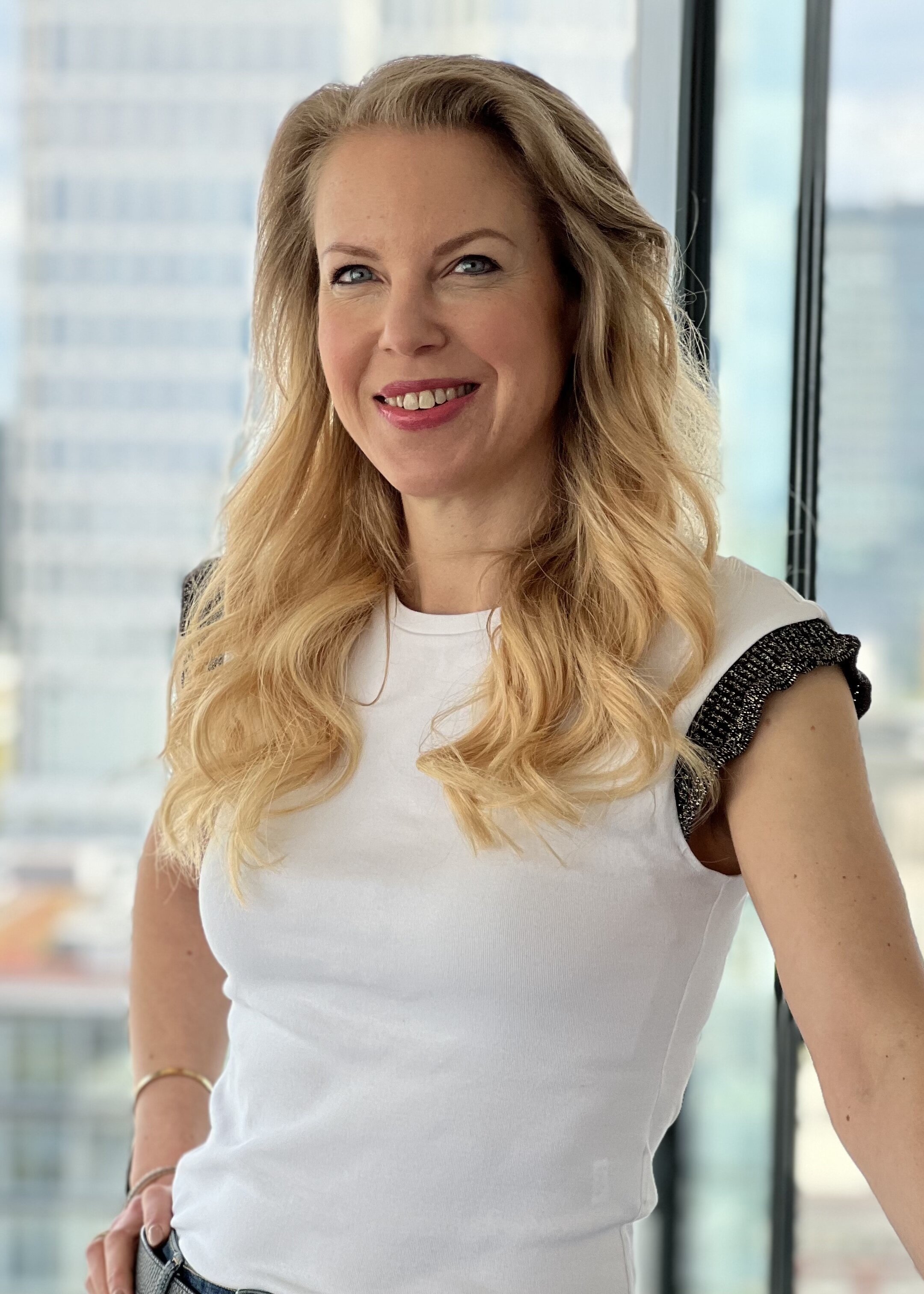 Viola Herrmann, Redakteurin, Porträt mit strahlendem Lächeln und direktem Blick – schulterlange goldblonde Haare mit Wellen, blaue Augen, leuchtend pinker Lippenstift, weißes T-Shirt, schwarzer Gürtel mit goldenem Schnalle, schwarzes Armband, urbaner Hintergrund mit Skyline, professionell und selbstbewusst, Erfolg und Zugänglichkeit