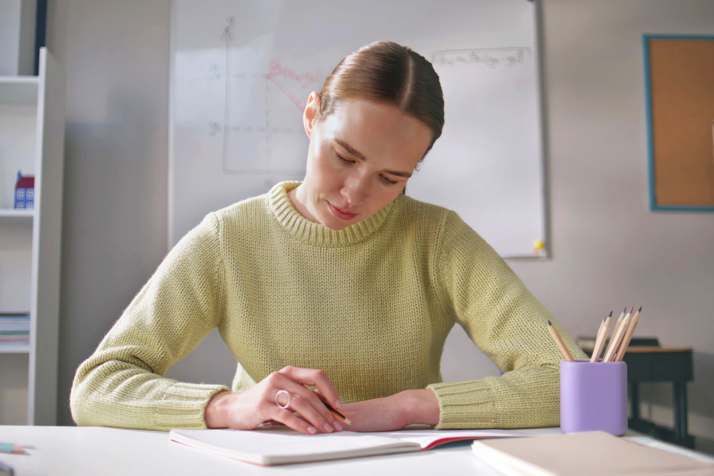 Frau mit braunem Haar in gelbem Pullover arbeitet konzentriert am Notizbuch mit Buntstiften, weiße Arbeitsumgebung mit Whiteboard und Regalen, Unterrichtsplanung