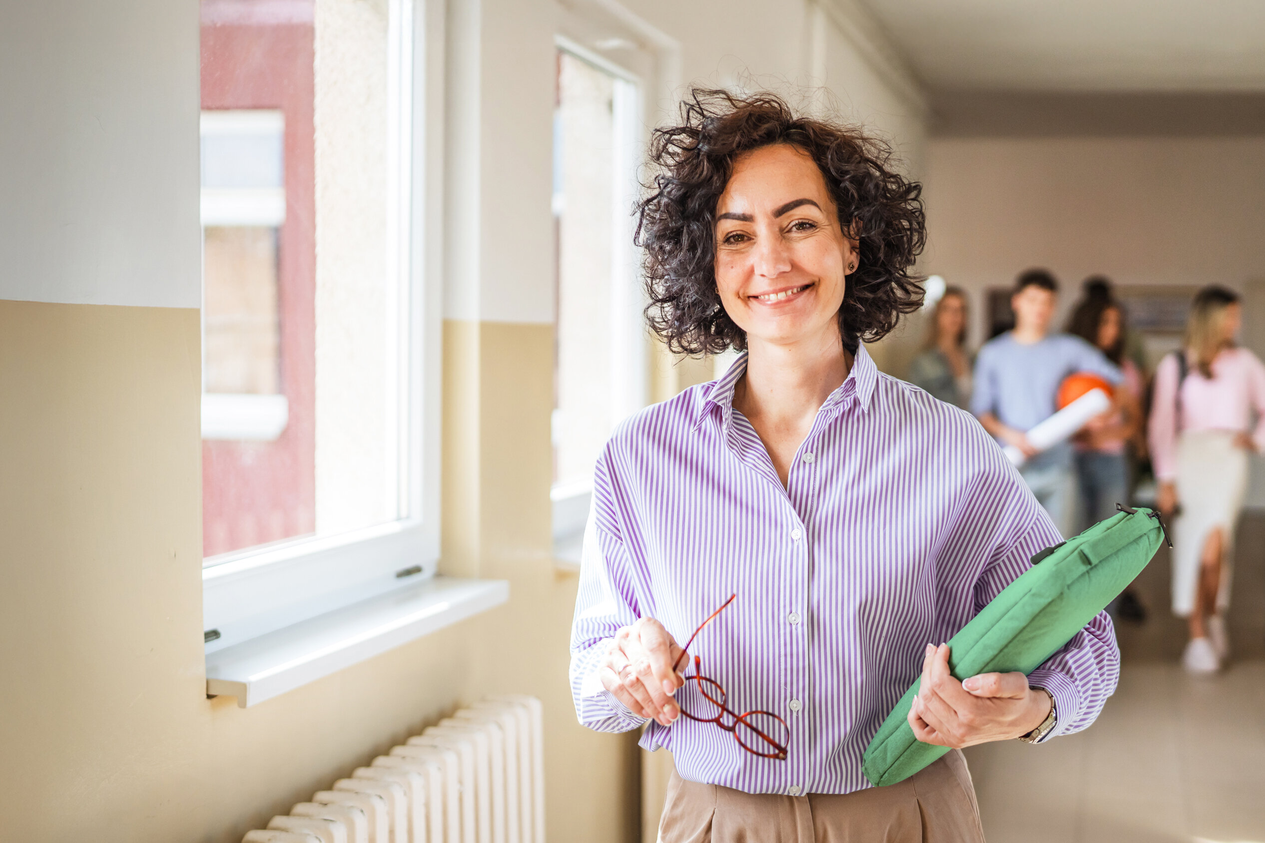 Lehrerin mit grünem Ordner und Brille im Schulflur mit Schüler:innen, moderne Schulumgebung, digitale Medienkompetenz