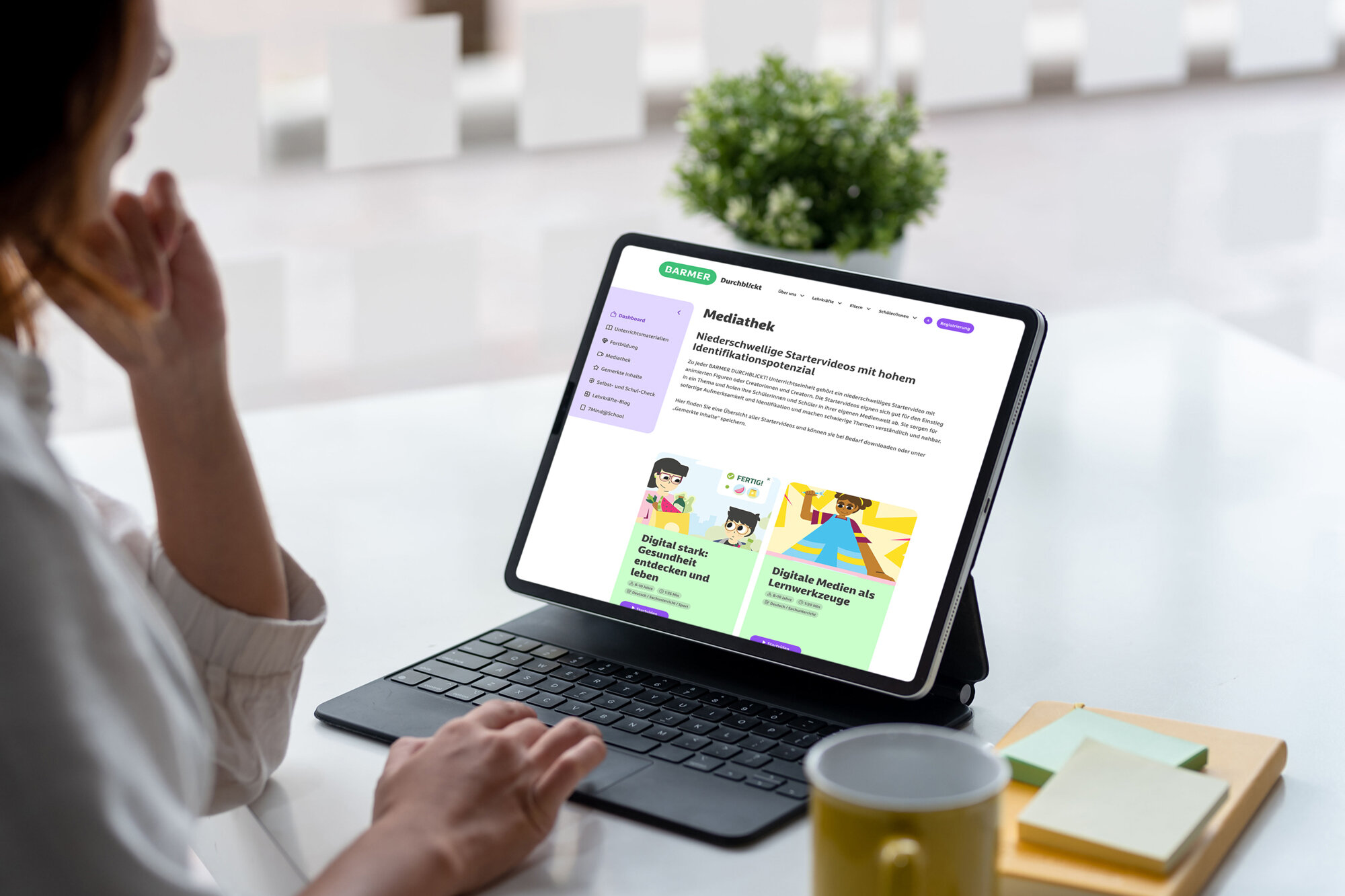 Mittelalte Lehrkraft mit welligem dunkelbraunem Haar in cremefarbenem Pullover navigiert konzentriert durch die BARMER Mediathek auf Tablet mit Detachable-Tastatur – Zugriff auf digitale Gesundheitsinhalte und Lernressourcen