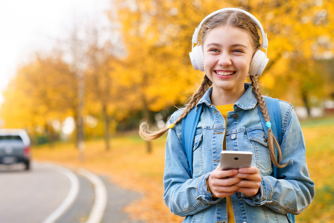 Ein lächelndes Mädchen mit Kopfhörern geht im Herbst an einer Strasse entlang. Sie hat ein Smartphone in der Hand.