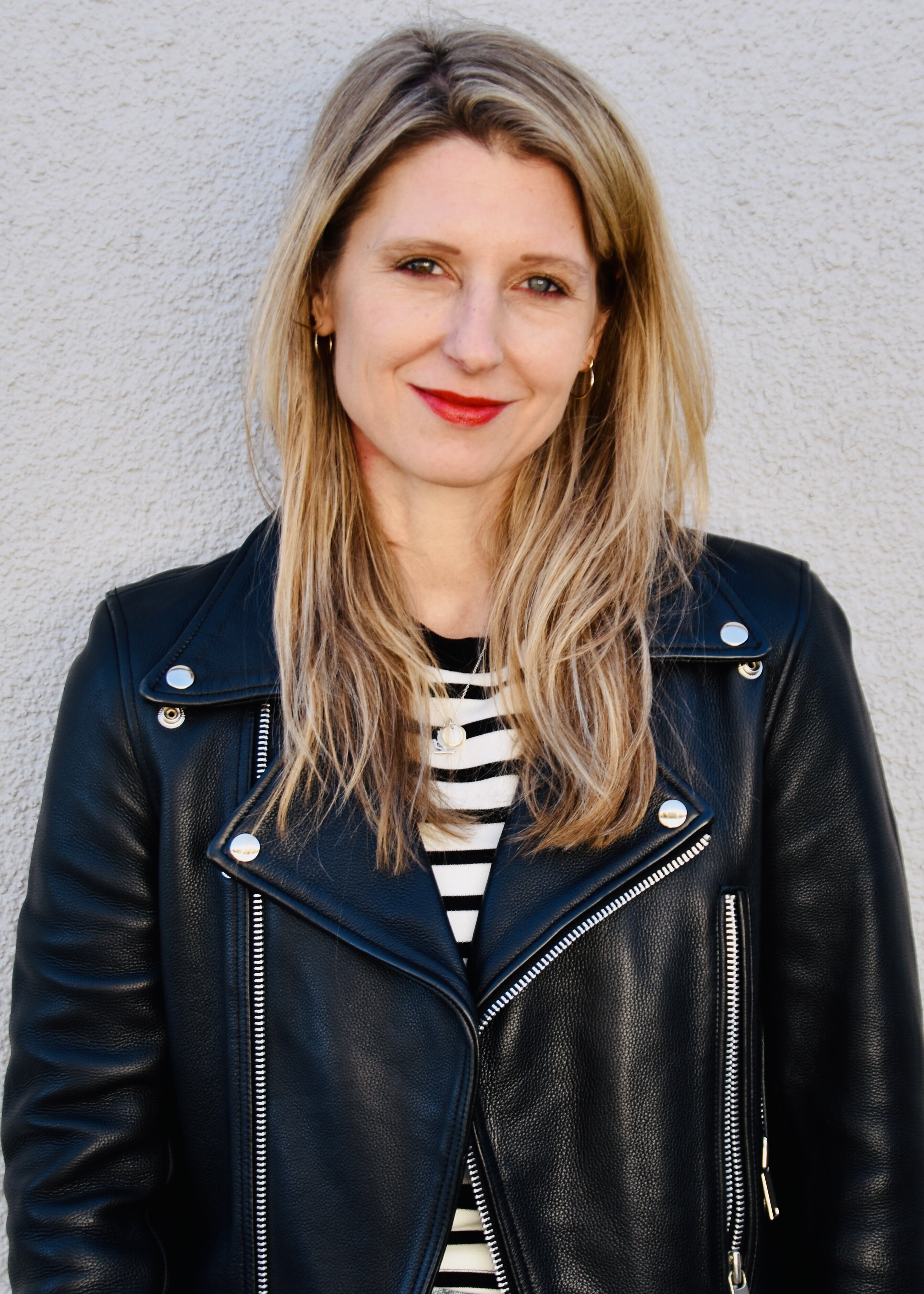 Laura Fröhlich, Redakteurin, Porträt mit warmem Lächeln und direktem Blick – schulterlange braune Haare mit Highlights, roter Lippenstift, schwarze Lederjacke über Streifenshirt, professionell und nahbar, Vertrauen und Expertise