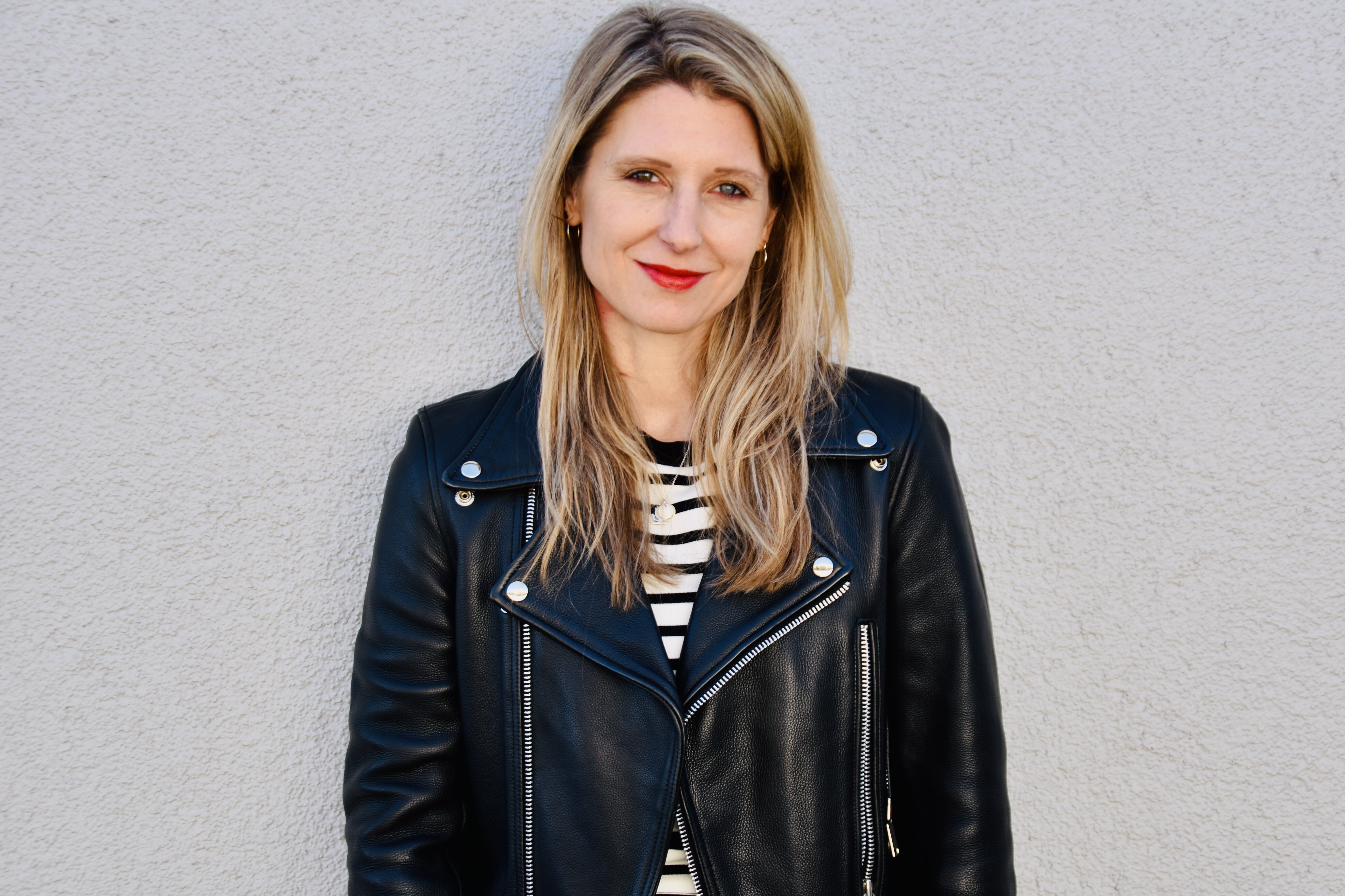 Laura Fröhlich, Redakteurin, Porträt mit warmem Lächeln und direktem Blick – schulterlange braune Haare mit Highlights, roter Lippenstift, schwarze Lederjacke über Streifenshirt, professionell und nahbar, Vertrauen und Expertise