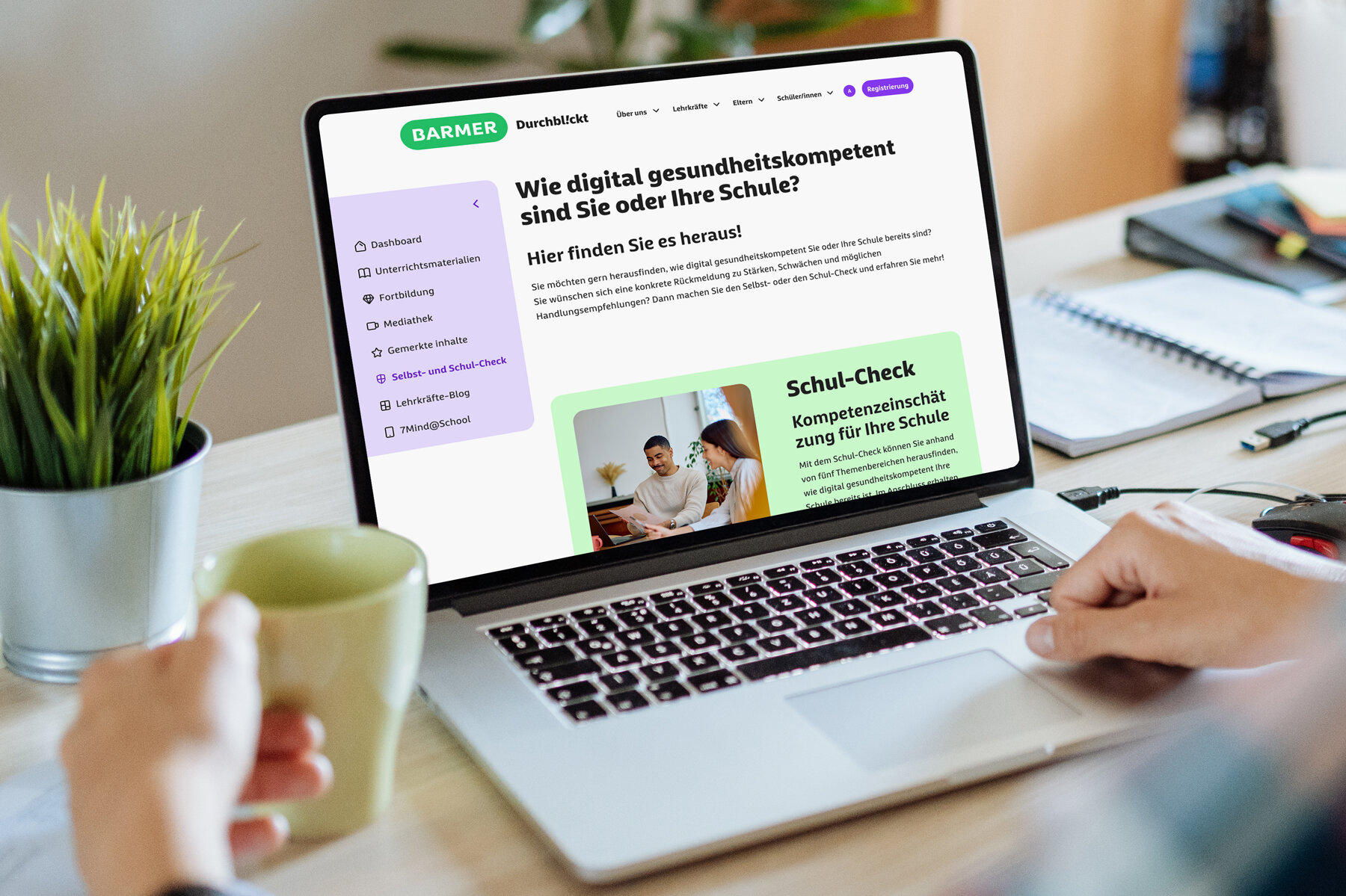 Lehrerin am Holzschreibtisch mit Laptop zeigt BARMER Durchblickt Schul-Check zur digitalen Gesundheitskompetenz, grüne Buttons, moderne Schulumgebung