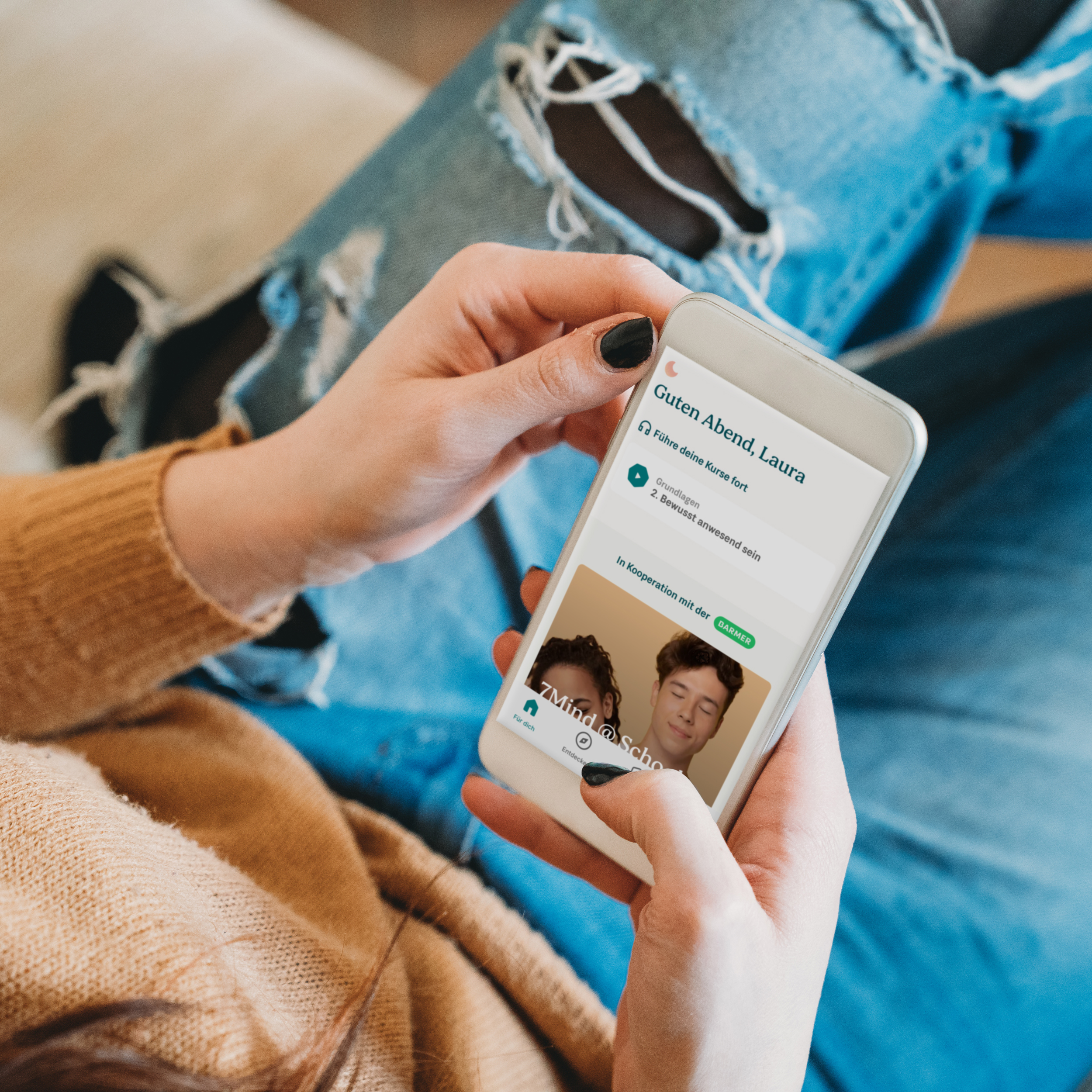 Junge Frau mit beiger Strickjacke und blauer Jeans hält weißes Smartphone mit 7Mind-Kurs-App – „Guten Abend, Laura", Kurse fortsetzen, digitale Gesundheitskompetenz, Stressabbau, In Kooperation mit 7Mind