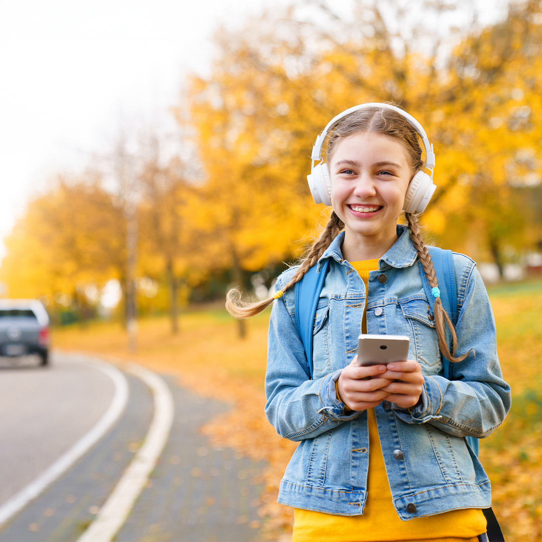 Ein lächelndes Mädchen mit Kopfhörern geht im Herbst an einer Strasse entlang. Sie hat ein Smartphone in der Hand. DGK_Artikel-17_Use-of-media.jpg