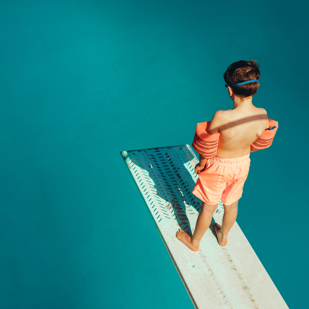 Junge mit Schwimmflügeln steht auf Sprungbrett im Schwimmbad und schaut ins Becken. DGK_Artikel-06_Challenges.jpg