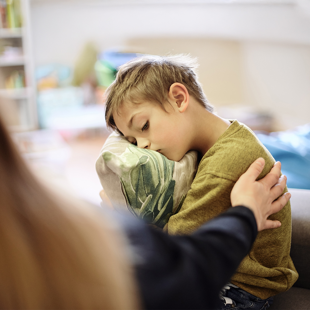 Ein Junge sitzt traurig auf einem Sofa und hält ein Kissen im Arm. Eine erwachsene Person legt ihm tröstend die Hand auf die Schulter. DGK_Artikel-06.01_eltern_ADHS.jpg