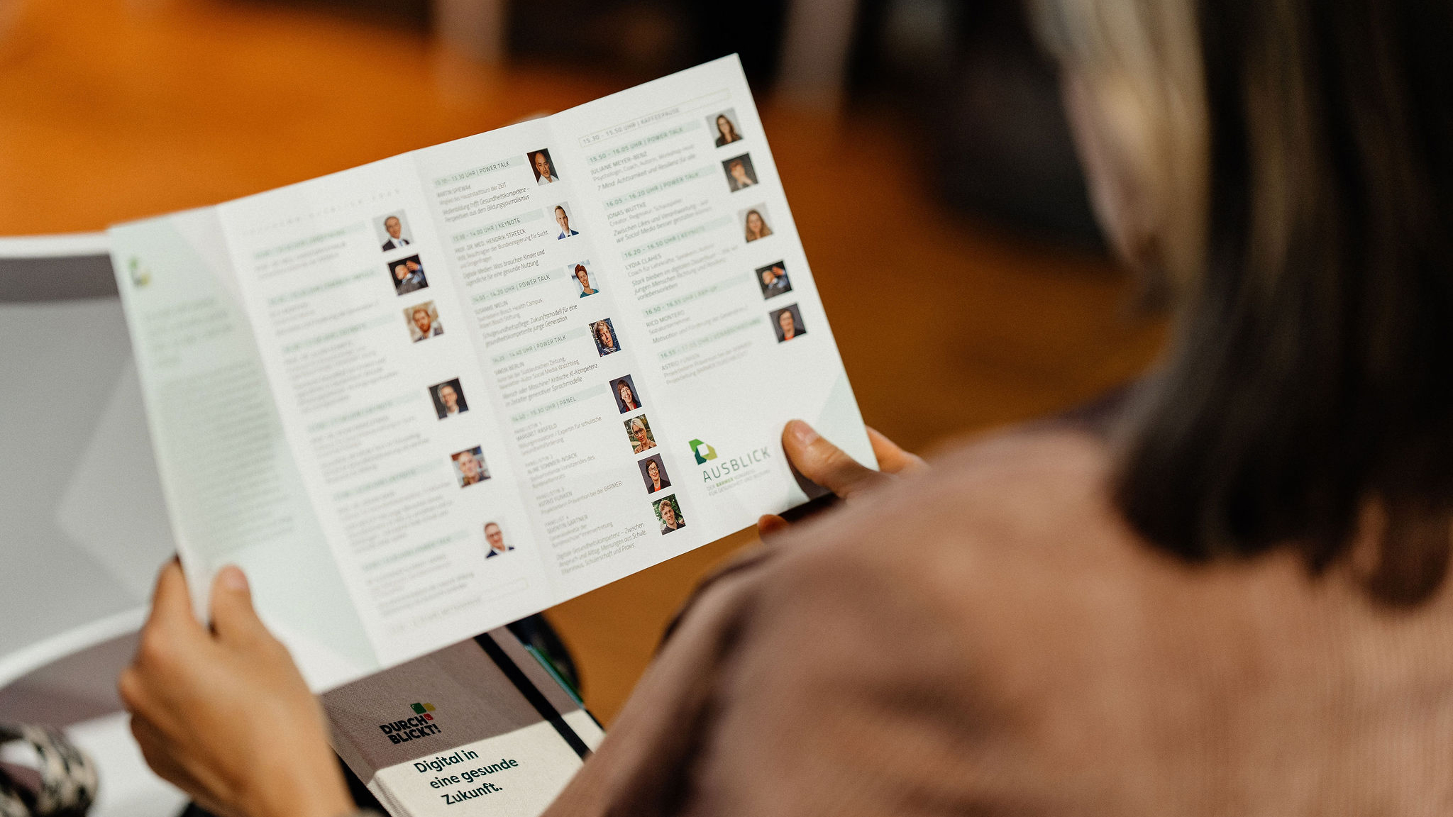 Person hält aufgeschlagene AUSBLICK-Programmbroschüre mit Referent:innen-Porträts, Zeitplänen, grünen Highlights, Vortragsreihe für Lehrkräfte