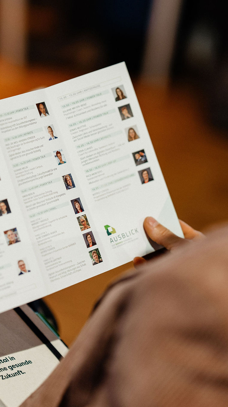 Hände halten aufgeschlagene Broschüre mit Vortragsprogramm, Referent:innen-Fotos, warmer Holzhintergrund, Digitale Gesundheit Vortragsreihe
