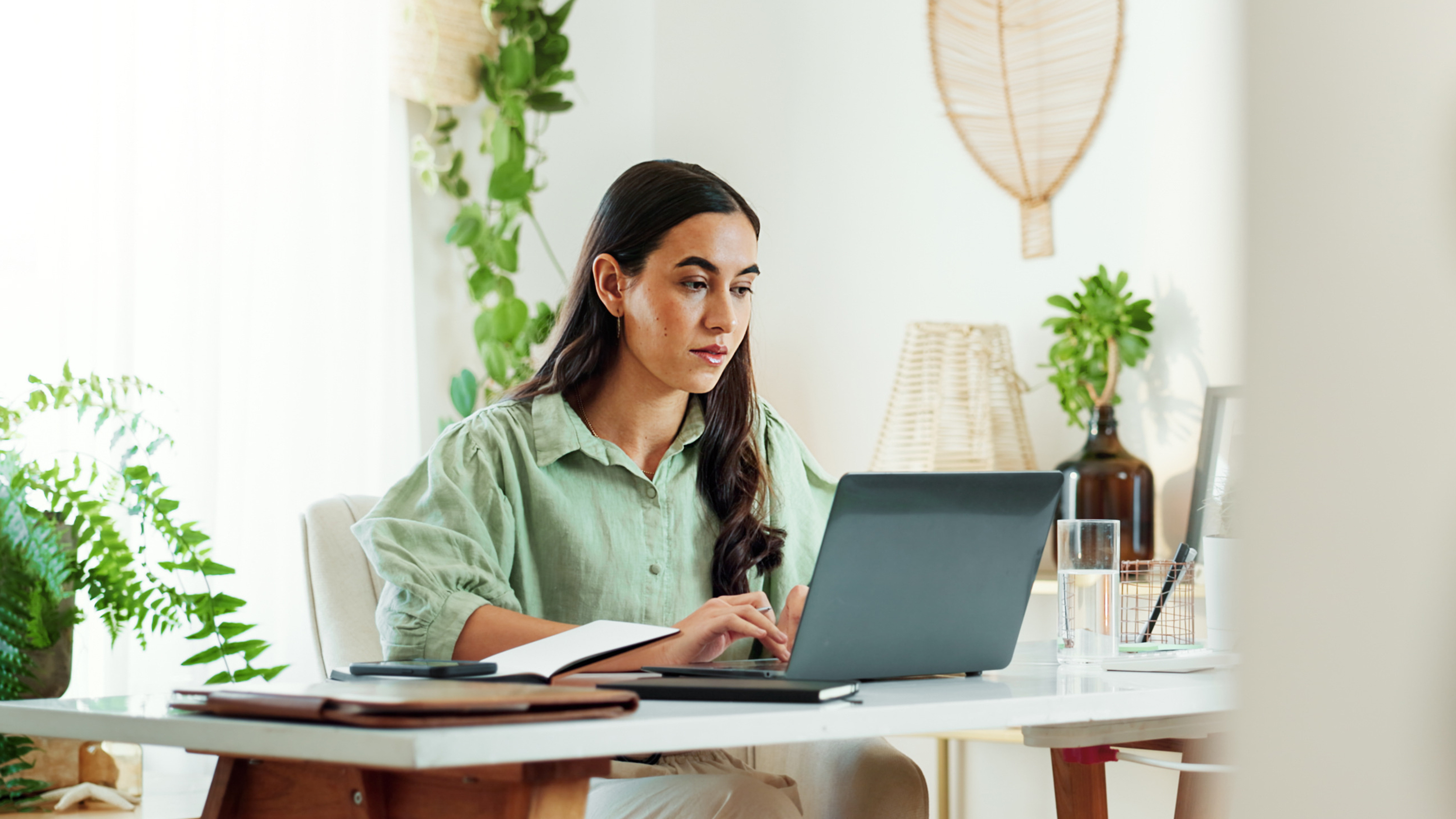 Frau mit schwarzen Haaren arbeitet konzentriert am Laptop mit Notizbuch, grünes Hemd, pflanzenreicher Workspace