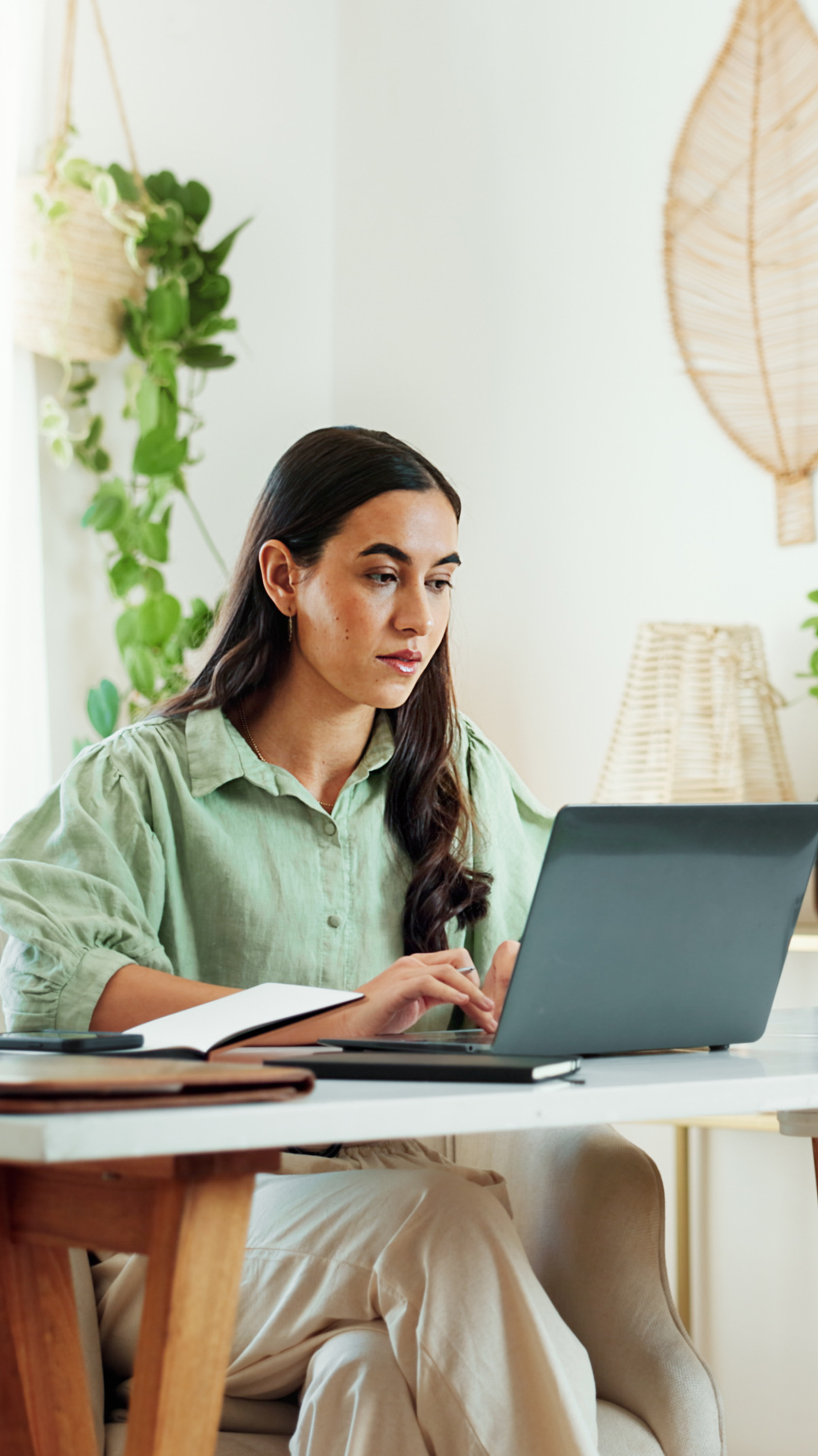 Frau am Laptop mit Notizbuch, helles Home-Office mit Pflanzen, ruhige Arbeitsatmosphäre