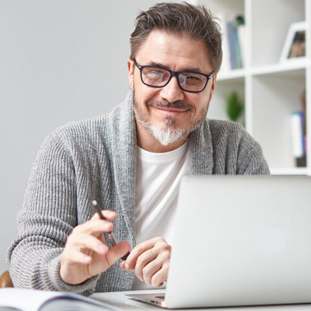 Mann mit Brille und grauer Strickjacke sitzt vor Laptop und blickt lächelnd in die Kamera