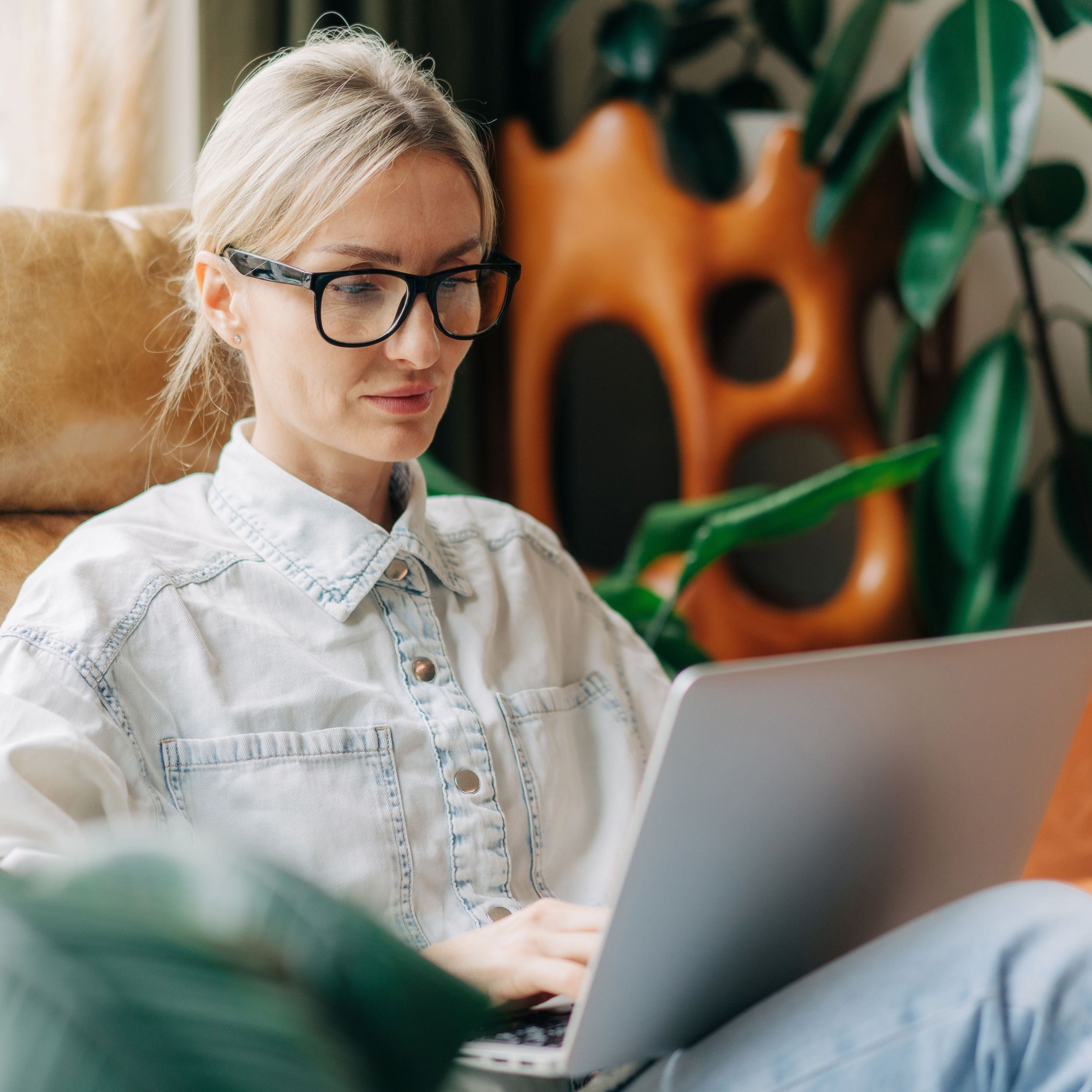 Blonde Frau mit Brille in hellblauer Denim-Bluse arbeitet konzentriert am Laptop, grüne Pflanzen und orange Kunstskulptur im Hintergrund, moderne Arbeitsumgebung