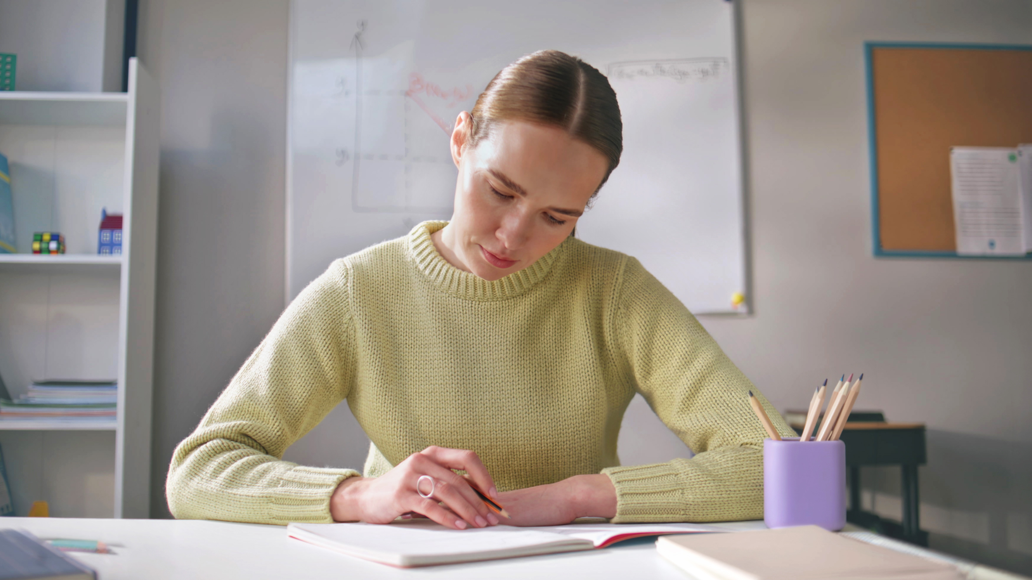 Frau mit braunem Haar in gelbem Pullover arbeitet konzentriert am Notizbuch mit Buntstiften, weiße Arbeitsumgebung mit Whiteboard und Regalen, Unterrichtsplanung