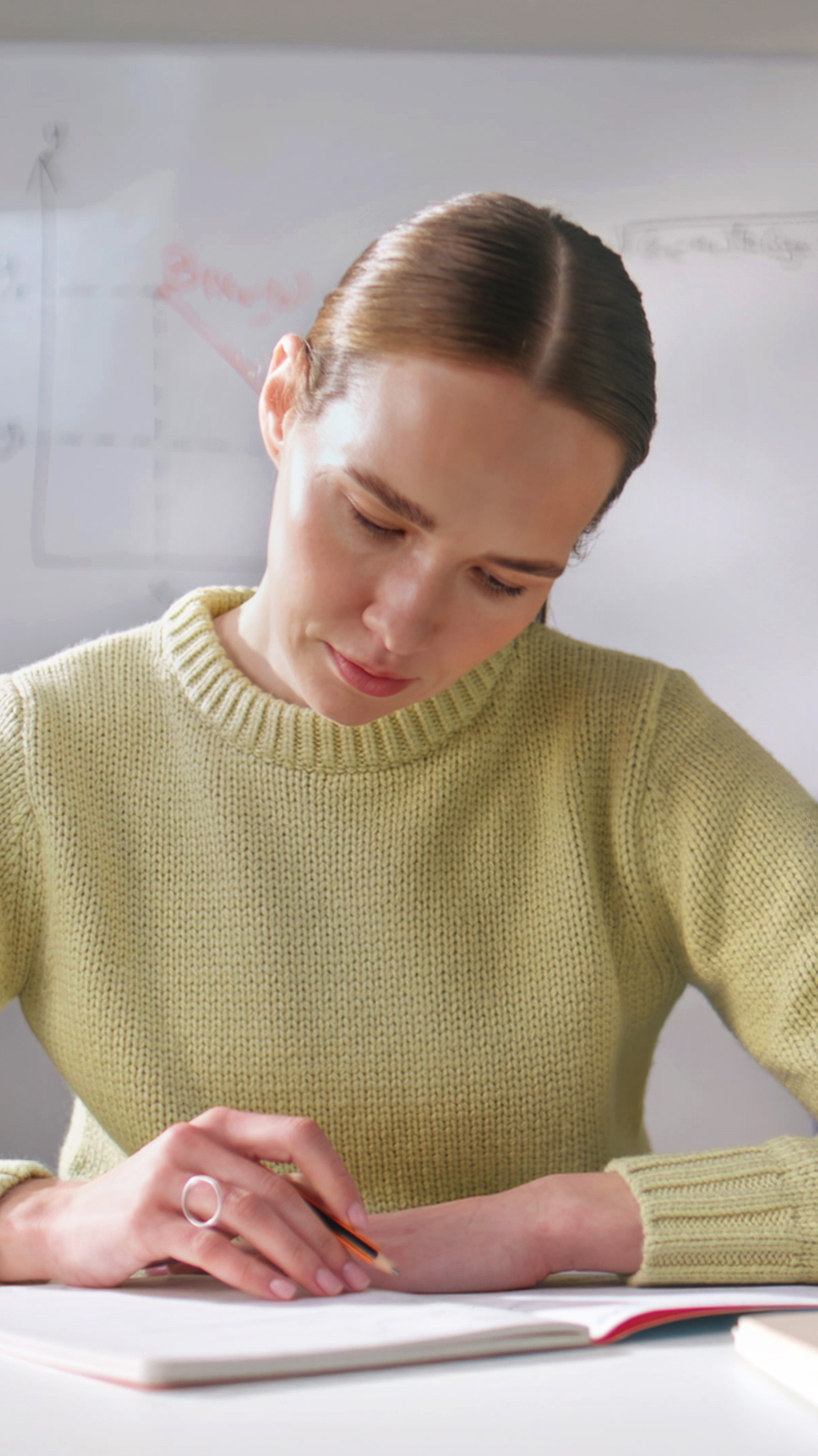 Frau mit braunem Haar in gelbem Pullover schreibt konzentriert auf Arbeitsblatt, Stift in Hand, heller Schreibtisch, Unterrichtsplanung und Vorbereitung