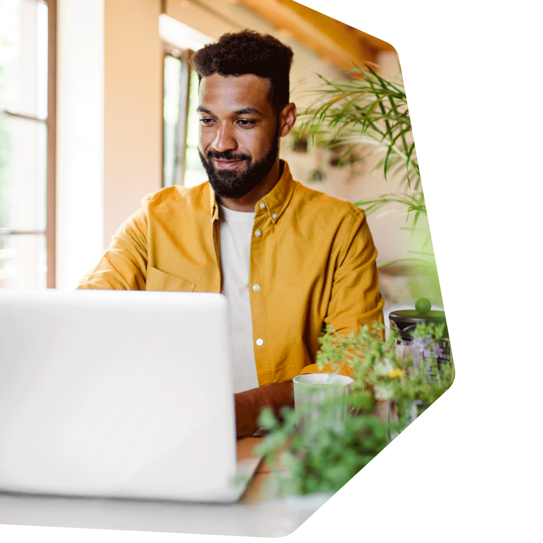 Mann mit dunklem Vollbart in gelbem Hemd arbeitet konzentriert am Laptop, grüne Pflanzen und Fenster im Hintergrund, moderne Homeoffice-Umgebung