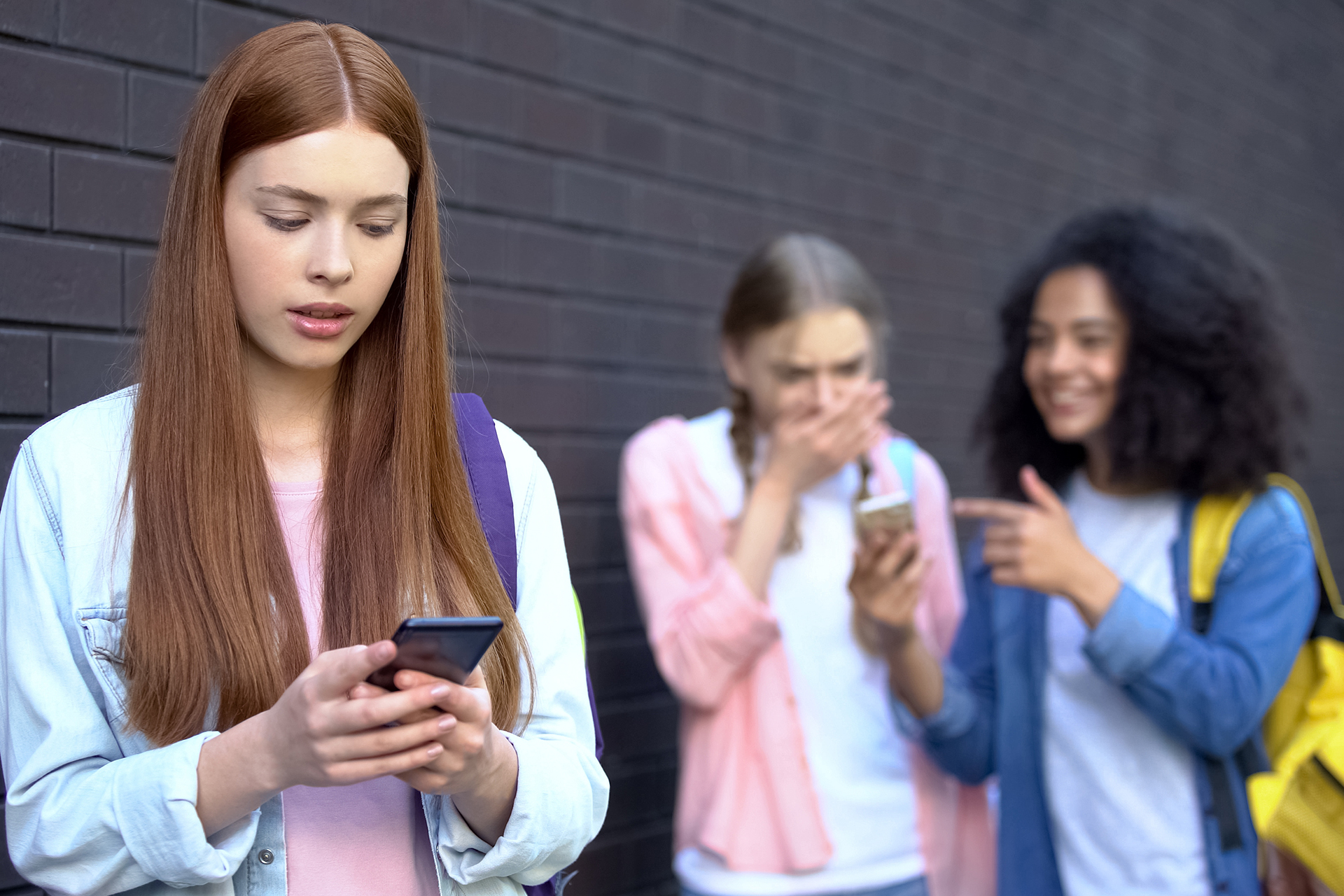 Mädchen sieht verunsichert auf Smartphone. Im Hintergrund lachen Mädchen die auf ein Smartphone schauen
