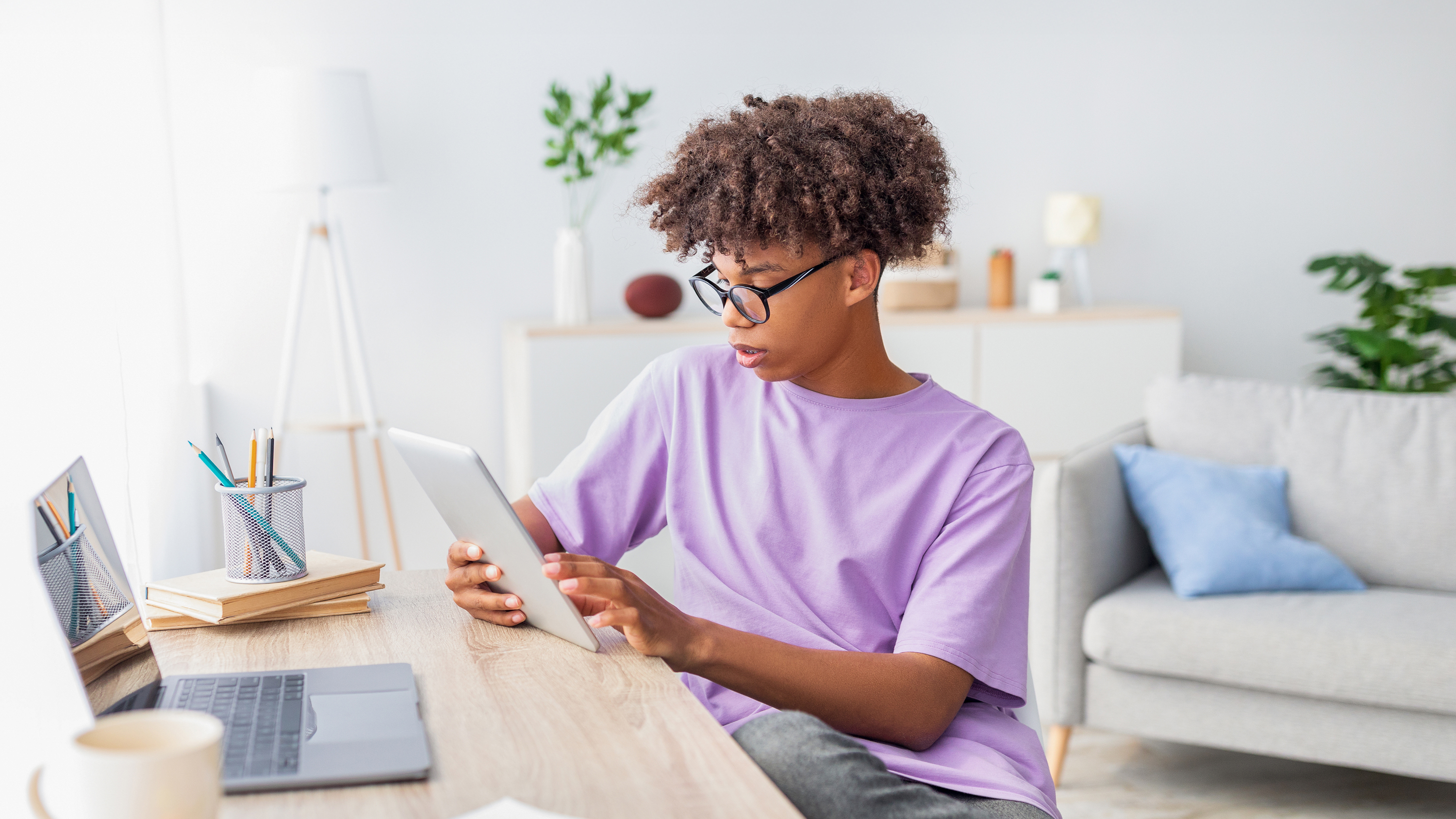 Jugendlicher mit schwarzer Brille und Afro-Frisur am Schreibtisch, Tablet in Händen, lila Shirt, digitales Lernen zuhause