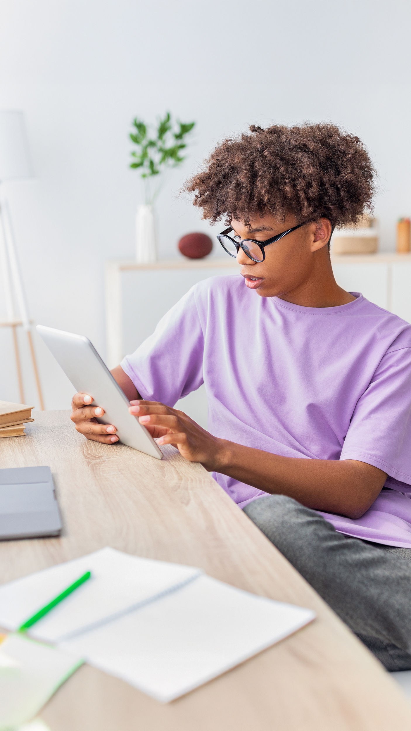Jugendlicher mit schwarzer Brille und Afro-Frisur am Schreibtisch, Tablet in Händen, lila Shirt, digitales Lernen zuhause