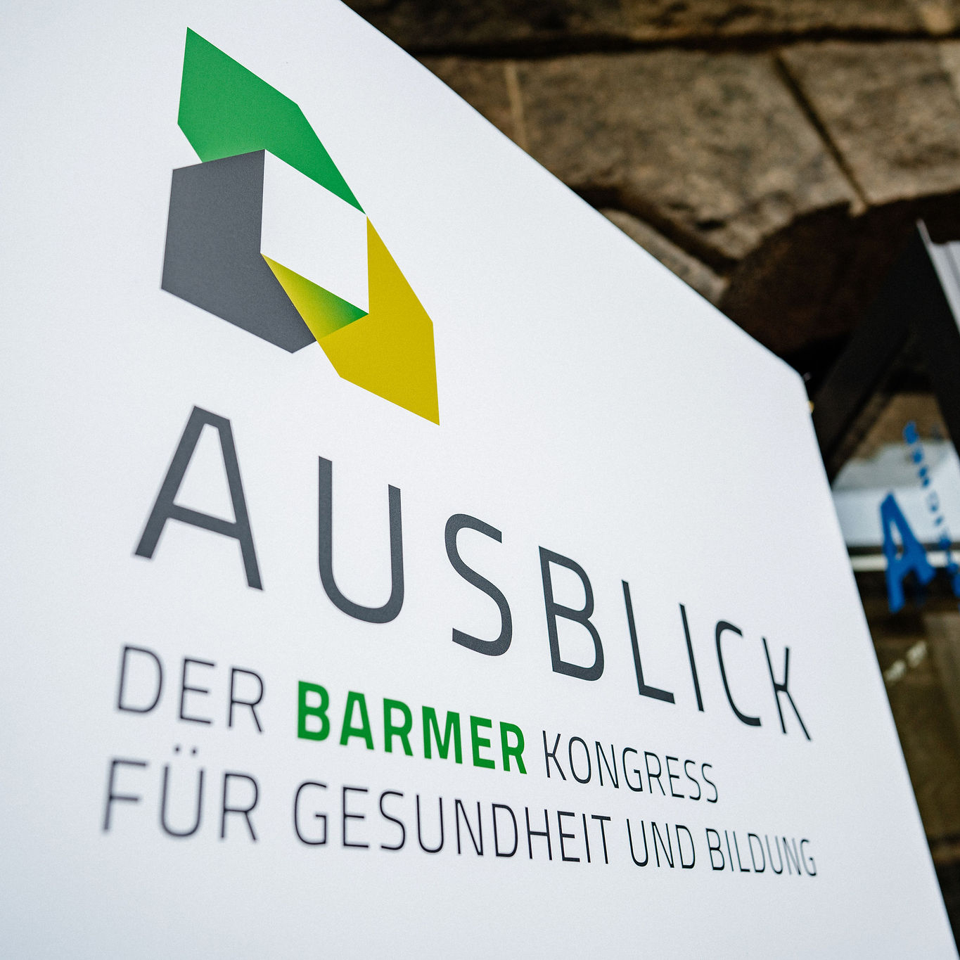 Professionelle Außenschild des BARMER Kongress "Ausblick" mit modernem geometrischem Logo in Grün und Gelb auf weißem Hintergrund – Konferenz für Gesundheit und Bildung vor Steinarchitektur