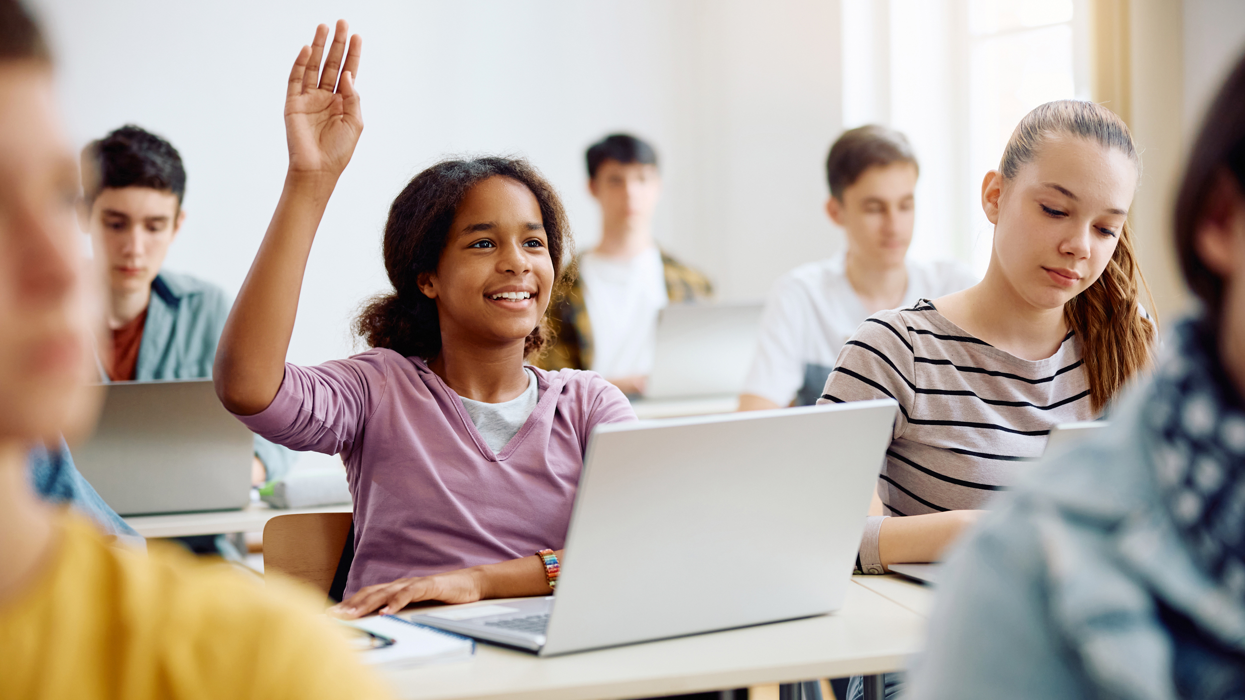 Diverse Schüler arbeiten aktiv mit Laptops im modernen Klassenzimmer – Schüler mit violettem Shirt meldet sich, Schülerin mit gestreiftem Shirt konzentriert auf Bildschirm