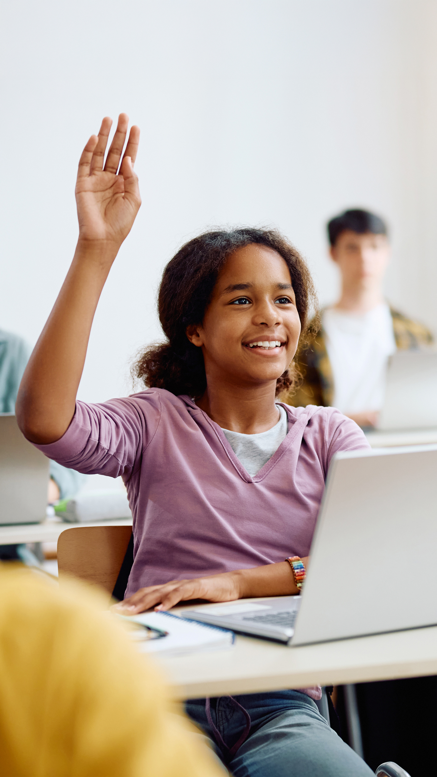 Schülerin mit Pferdeschwanz meldet sich aktiv mit gehobener Hand im hellen Klassenzimmer mit Laptop und Schulheften